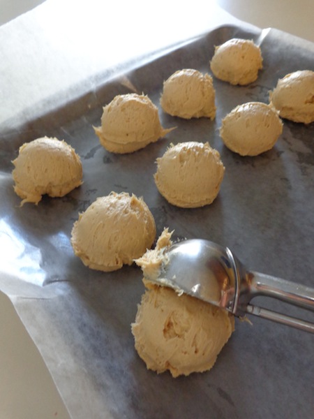 sheet pan covered with wax paper and topped with round scoops of peanut butter cheesecake balls - one scoop is being scooped out of medium scoop